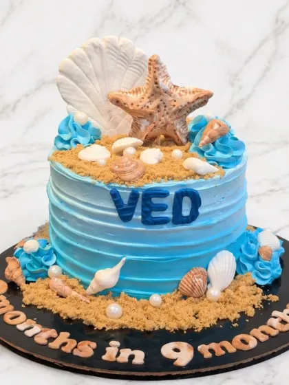 An ocean-themed cake celebrating "9 months in, 9 months out". The blue ombre cake is decorated with edible sand and detailed fondant seashells.