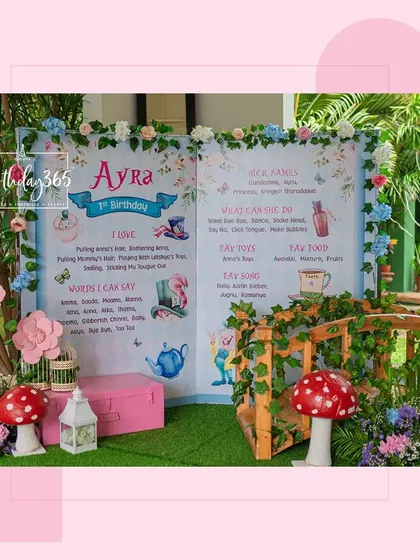 A creative 'Alice in Wonderland' themed backdrop for a first birthday. Designed like an open storybook, the display shares fun facts about the birthday girl, Ayra, surrounded by whimsical illustrations and floral decor.