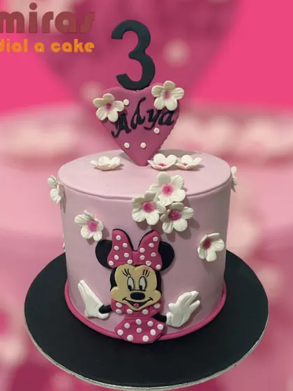 A pretty pink Minnie Mouse cake for a third birthday. The cake is decorated with small white flowers and a heart-shaped topper, featuring the charming and iconic Minnie.