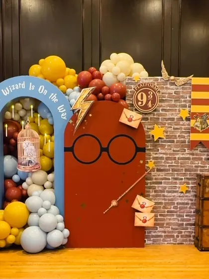 "A Little Wizard is On the Way." A magical Harry Potter-themed baby shower setup, complete with a Platform 9 3/4 brick wall, iconic glasses and scar, a golden snitch, and house color-themed balloons.