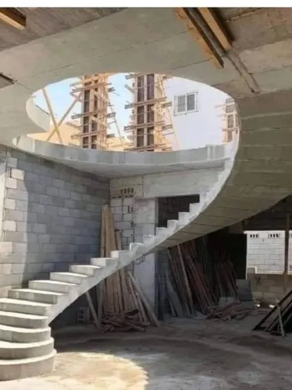 A stunning spiral staircase made of concrete, captured during the construction phase. The smooth, curving form and the oculus opening in the ceiling create a powerful architectural moment even in its raw state.