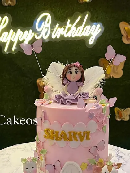 A beautiful fairy-themed cake set against a party backdrop with a 'Happy Birthday' neon sign. The cake features a fairy with white wings and is surrounded by butterflies.