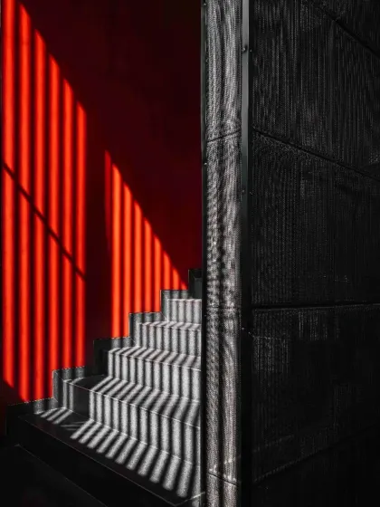 Light and shadow create a dynamic pattern on the staircase of Project 1374. The bold red wall contrasts with the dark, charred wood and the textured concrete, showcasing our approach to creating a rich, layered tapestry of materials.