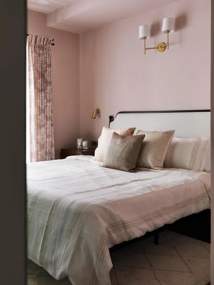 Another view of the dusty rose bedroom from the Madras Home. The soft color, paired with a simple metal frame bed and patterned curtains, creates a cozy and inviting atmosphere.