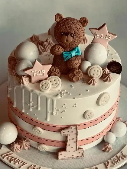 A lovely first birthday cake with a hand-sculpted teddy bear topper. The design includes delicate details like edible buttons, stars, and the baby's birth date for a personal touch.