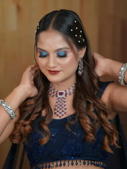 A close-up of a glamorous party look with bold blue glitter eyeshadow and a deep red lip. The hair is adorned with scattered pearls for a touch of elegance.