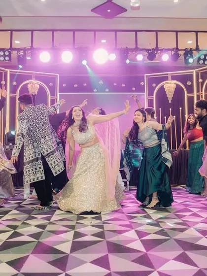A wide shot of a choreographed group dance at a Sangeet, with dramatic stage lighting and a checkered dance floor.