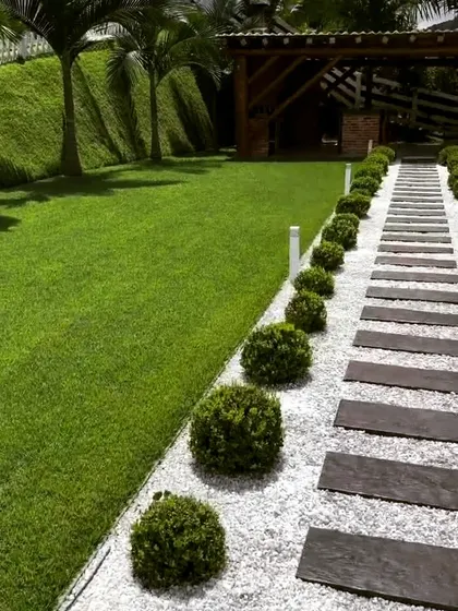 A garden path made of dark stone slabs set in a bed of white gravel, running alongside a lush lawn. The path is lined with neatly trimmed spherical shrubs, adding a formal, structured element to the landscape.