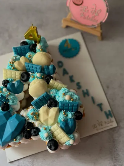 A stylish monogram cake in shades of blue and white for Dakshith's fourth birthday. This number 4 cake is loaded with toppings like blueberries, white and blue chocolate bars, and a geometric chocolate heart.
