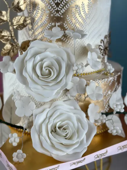 A symphony of white and gold to celebrate a beautiful love story. This elegant tiered cake features intricate textures, gold leaf details, and stunning handcrafted white sugar roses.