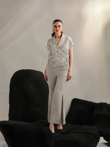 A full-length look featuring an embellished wrap top and flared trousers. The model stands atop black geometric blocks, creating a strong, architectural composition.