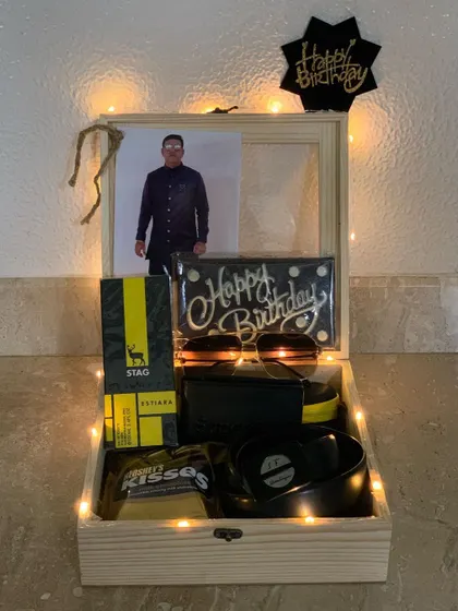 Celebrate him with a personalized wooden keepsake box. This birthday hamper includes a custom photo, a 'Happy Birthday' chocolate bar, stylish sunglasses, a wallet, and other treats, all beautifully arranged and lit with fairy lights.