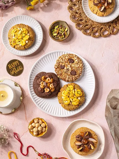 A top-down flat lay of assorted Rakhi cookies, styled with a candle and traditional rakhi on a soft pink background.