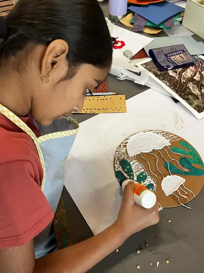 A close up view of a student meticulously adding details to her collagraph plate. She is using glue and various textured materials to create a scene with jellyfish, which will then be inked and printed.