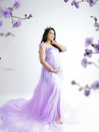 A whimsical and dreamy portrait. The mom-to-be, wearing a tiara and a lavender gown, is framed by delicate purple flowers.