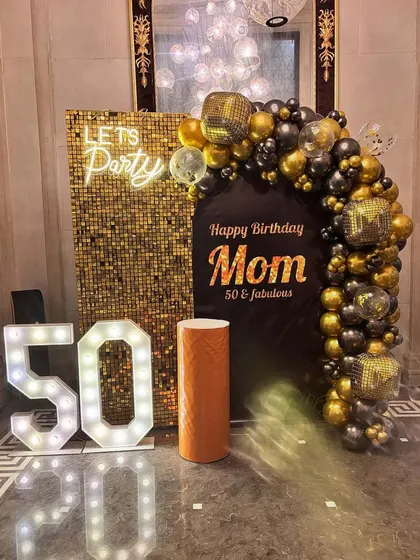 A glamorous 50th birthday decoration for 'Mom'. The setup combines a gold sequin panel with a custom black backdrop, a 'Lets Party' neon sign, and a black and gold balloon arrangement.