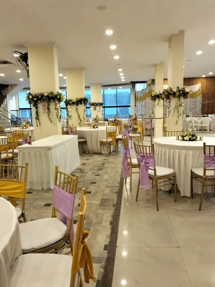 An overview of an anniversary celebration hall we decorated. The space features round tables with white linens, chairs accented with lavender sashes, and floral arrangements on the pillars, creating a cohesive and elegant look.