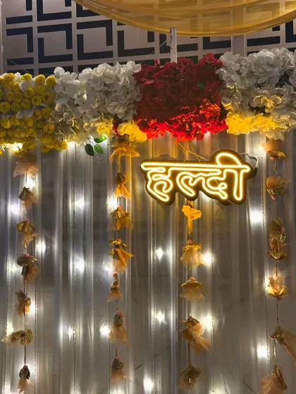 A close-up of the 'Haldi' neon sign against a white curtain with fairy lights. The sign is topped with a beautiful arrangement of red, white, and yellow flowers.