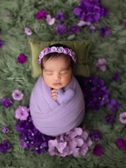 A close-up of a newborn wrapped in purple, holding a tiny felt heart. This shot emphasizes the baby's peaceful expression and the beautiful floral arrangement.