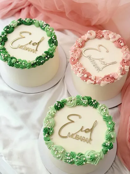 A set of three elegant mini cakes for Eid. Each cake is topped with a gold "Eid Mubarak" plaque and a wreath of piped buttercream in shades of green and pink.