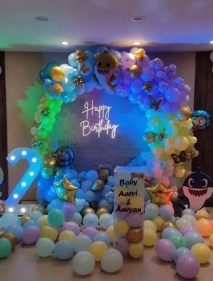 A Baby Shark themed second birthday party. The decor includes a circular balloon arch, character cutouts, and a '2' marquee light, all illuminated with colorful lighting.