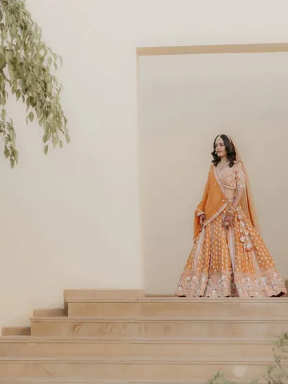 A full-length bridal portrait showcasing the magnificent orange lehenga against a minimalist architectural backdrop. This shot is all about grace and style.