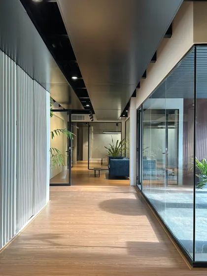 A primary office corridor lined with floor-to-ceiling glass on one side and textured paneling on the other. This design floods the circulation path with natural light and offers constant visual access to the outdoor green spaces.
