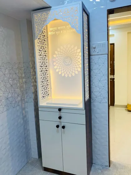 A modern mandir design with a white frame and dark wood sides. The backlit panel features an 'Om' mandala, and the unit includes drawers and cabinets for storage.