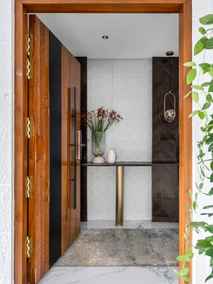 The entrance to Dr. Harsha's home offers a glimpse of the minimalist luxury within. A custom carved wooden door opens to a foyer with clean lines, a sleek console table, and contrasting dark wood paneling.