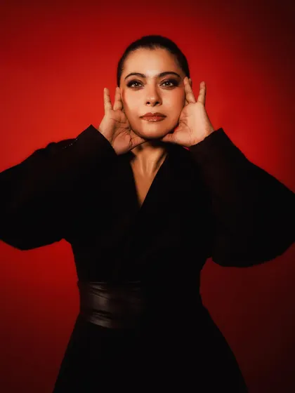 Kajol in a dramatic all-black wrap dress with a leather belt, set against a bold red background for the 'Do Patti' promotions.