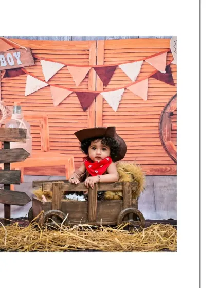 A portrait of a baby boy in a wooden cart as part of a 'Cowboy' theme. The rustic props and western-style outfit make for a truly unique milestone photograph.