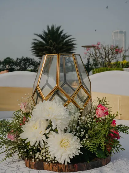 A modern and elegant centerpiece combining a geometric gold lantern with fresh white chrysanthemums and greenery on a rustic wood slice base.