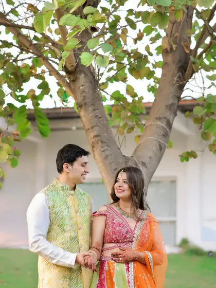A sweet, candid moment during a pre-wedding shoot. The couple shares a look under a leafy tree, their colorful outfits popping against the natural background.