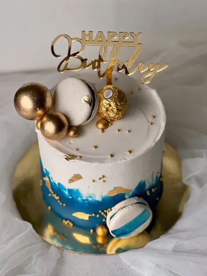 A stylish and masculine design perfect for him. This rich vanilla buttercream cake features a blue watercolor effect, gold leaf, macarons, and a Ferrero Rocher topper.