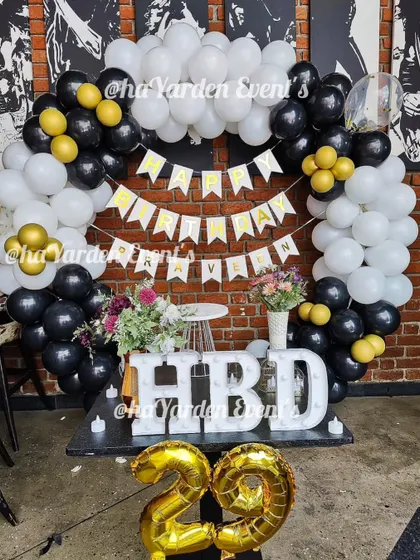 A fun and festive 29th birthday party. The black, white, and gold balloon arch is paired with large 'HBD' marquee letters and foil '29' balloons.