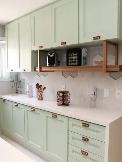 A charming kitchen with sage green cabinets and copper handles. The hexagonal tile backsplash and open wooden shelf add a touch of rustic, farmhouse-style elegance.