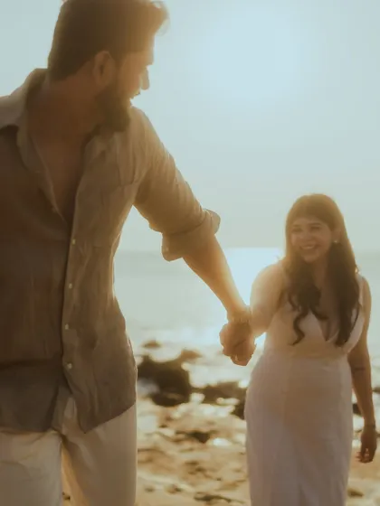 A sweet moment captured at sunset on the beach, with the groom leading the bride by the hand as she smiles at him. The golden hour light creates a warm and romantic atmosphere.