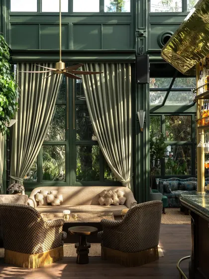 A sun-drenched seating area in the Lord Elgin Glasshouse. The design layers rich, patterned fabrics, fringed armchairs, and warm brass accents against the deep green of the structure to create a sense of exotic escapism.