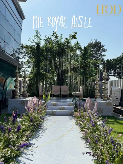 The "Royal Aisle" in its full glory. This outdoor wedding aisle combines a clean white path with lush floral borders and a stunning natural tree backdrop, perfect for a daytime garden wedding.