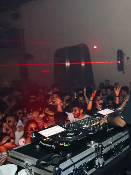 A view from behind the decks, showing the packed crowd and laser beams cutting through the darkness.