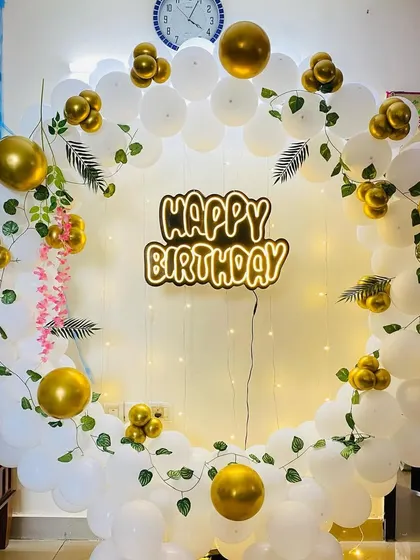 An elegant birthday balloon ring in white and gold, decorated with artificial vines and a custom neon sign.