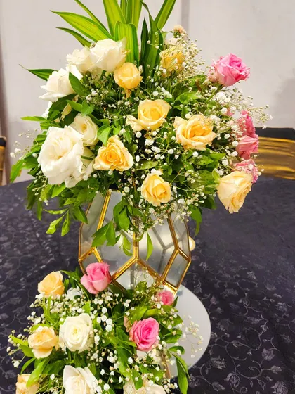 A beautiful floral arrangement in a geometric gold and glass vase. The bouquet features a soft palette of yellow, white, and pink roses, perfect for an elegant reception or gala dinner.