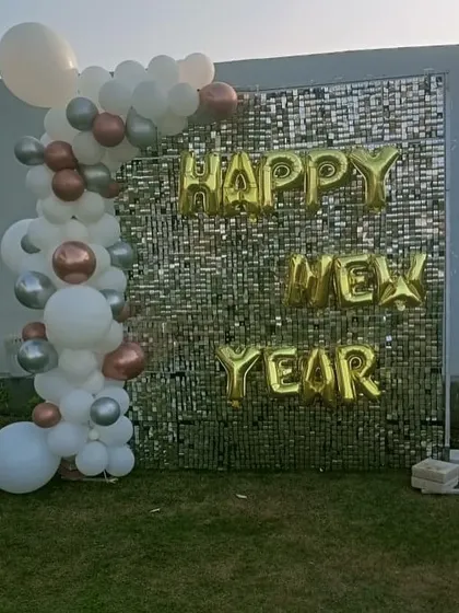 A festive 'Happy New Year' photo backdrop for a corporate party or event. A silver sequin wall is paired with a simple balloon garland and gold foil letters.