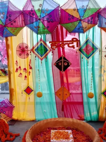 A vibrant Haldi ceremony space featuring a multi-colored backdrop with hanging kites and umbrellas. The 'urli' is filled with flower petals, ready for the rituals.