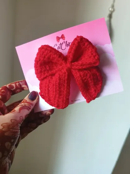Holding up a single red crochet sailor bow to show its size and texture. The quality of the handmade stitch is clear to see.