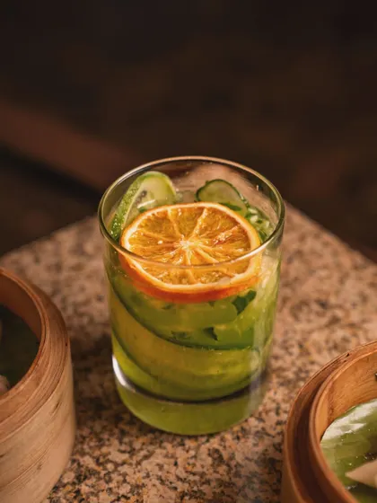 A refreshing green mocktail garnished with cucumber and a dehydrated orange slice. The moody lighting and presence of dim sum steamers in the background place the drink within an Asian dining context.