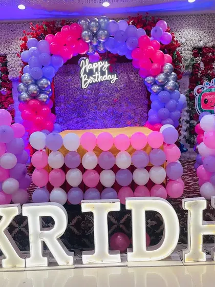 A full stage setup for a Cocomelon first birthday party. This grand decoration features a flower wall, a balloon arch, a custom table, and large light-up letters spelling the name 'KRIDHA'.