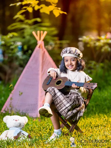 A little girl in a stylish plaid outfit plays her guitar while sitting in front of a teepee in a park. This outdoor setup is perfect for a fun and creative child photoshoot.
