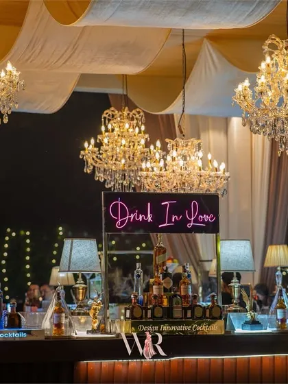 A stylish bar setup with a neon sign that reads "Drink In Love." The bar is lit by chandeliers and decorated with vintage-style table lamps, making it a chic and inviting focal point.