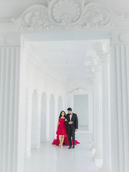 An expecting couple shares a quiet moment while walking through a majestic white corridor. This wide shot captures the grandeur of the architecture, framing them and her striking red dress in a truly cinematic style.